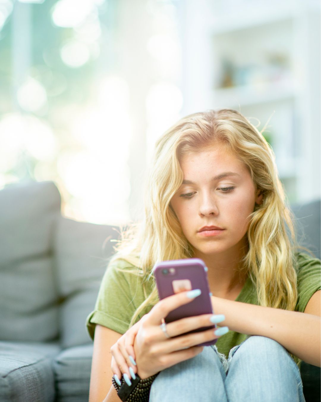 Young woman sitting on a couch, looking concerned while scrolling on her phone.